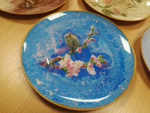 niebieski talerz ozdobiony metodą decoupage leżący na stole, rysunek na nim przedstawia szarego ptaszka siedzącego na gałązce z różowymi kwiatami