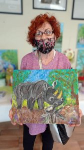 na pierwszym planie kobieta w maseczce i okularach, w różowej bluzce , trzyma przed sobą obraz malowany na płótnie przedstawiający nosorożca w tle obrazy wiszące na ścianie